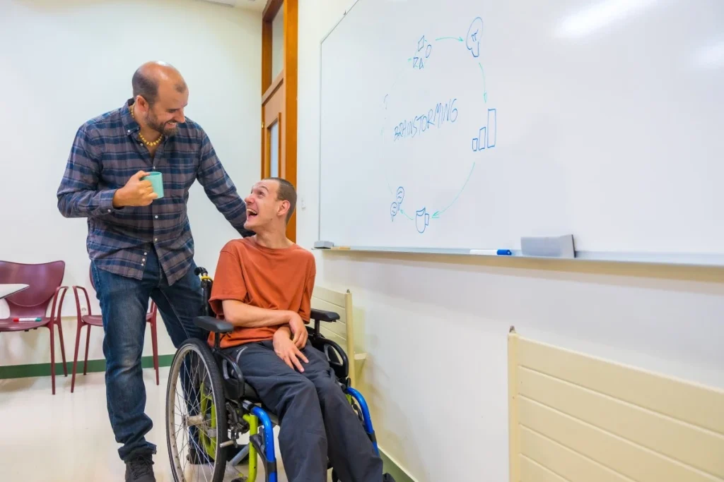 A disabled man and his teacher smiling at each other in front of a whiteboard